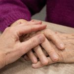 Två händer möts på ett bord - en äldre hand och en yngre hand. I bakgrunden syns en person i lila tröja. Bilden symboliserar mötet mellan generationer och värdighetsfull omsorg.